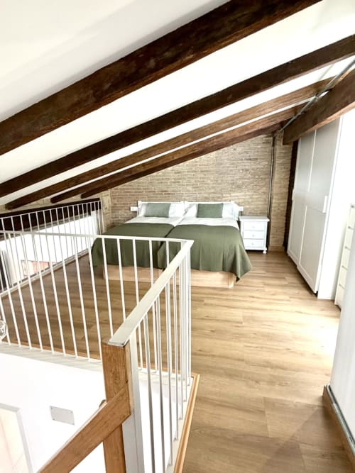 Dormitorio tipo loft con vigas de madera vista, ropa de cama verde, barandilla blanca, pared de ladrillo y suelo de madera en Stay Gest Valencia