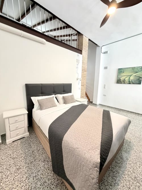 Habitación moderna en loft Stay Gest Valencia con cama queen, cabecero oscuro, mesilla blanca, ventilador de techo y ladrillo visto