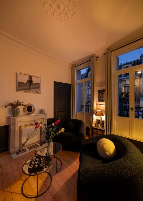 Elegant living room in Epernay apartment with black armchair, fireplace, city views through tall windows, and warm ambient