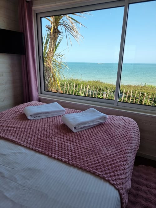 Chambre en bord de mer du gîte Bayeuzen avec vue sur l'océan, serviettes blanches sur le dessus-de-lit mauve, palmier et océan visibles