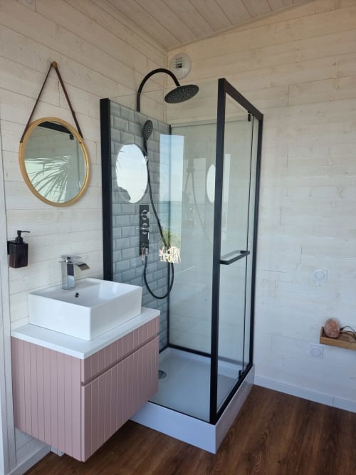 Salle de bain moderne du gîte Bayeuzen avec carrelage blanc, meuble rose, paroi de douche noire et miroir ovale
