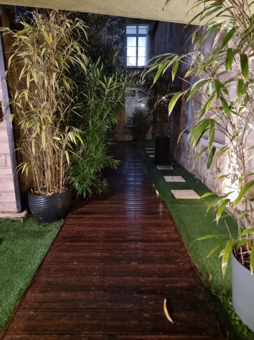 Passage verdoyant du jardin de la villa Le Jardin à Bayeux avec bambous, terrasse en bois et gazon synthétique