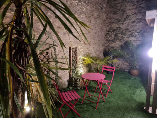 Coin salon jardin cosy à la villa Le Jardin de Bayeuzen avec chaises bistrot roses, table, plantes tropicales et murs en pierre rustique