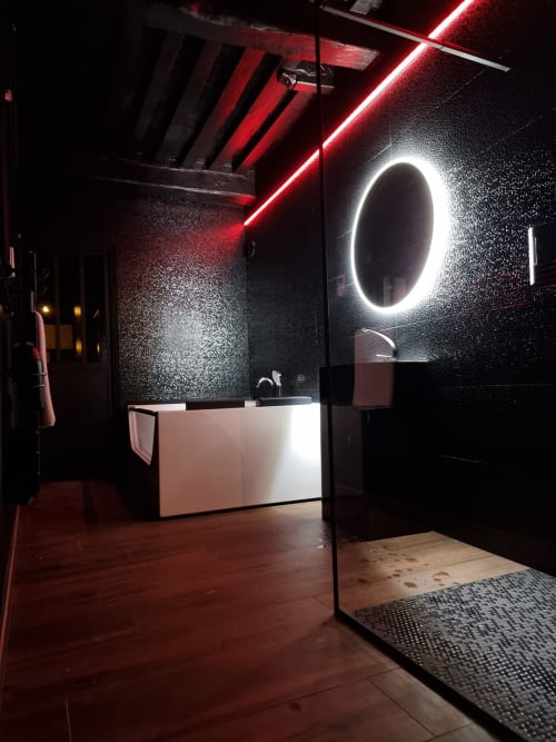 Salle de bain moderne avec miroir circulaire lumineux, éclairage néon rouge au plafond, murs texturés sombres et meuble vasque blanc à Bayeuzen