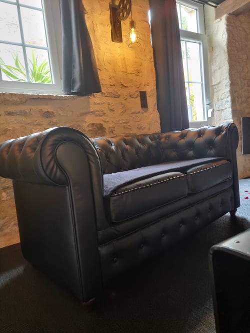 Canapé Chesterfield en cuir noir à la villa Le Jardin de la Chambre d'hôtes Bayeuzen, Bayeux, avec murs en pierre apparente et vue sur le jardin