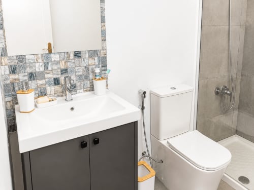 Baño moderno en apartamento de Tenerife con lavabo blanco, tocador oscuro, azulejos con patrón, inodoro y ducha a ras de suelo