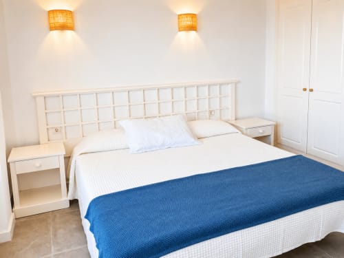 Luminoso dormitorio de apartamento en Tenerife con cabecero blanco de rejilla, cortina azul en la cama, mesillas a juego y vistas al mar
