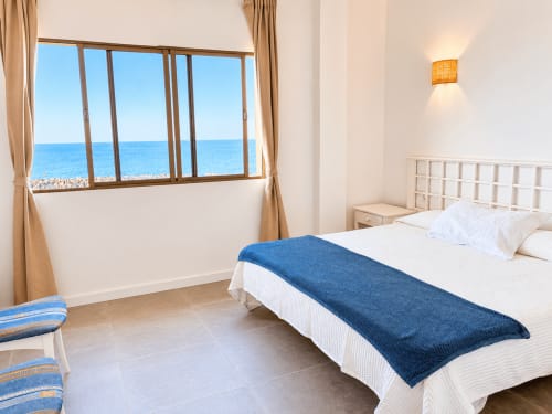 Dormitorio luminoso con vistas al mar en apartamento de Tenerife, océano azul visible por grandes ventanales, cama blanca con camino azul, y