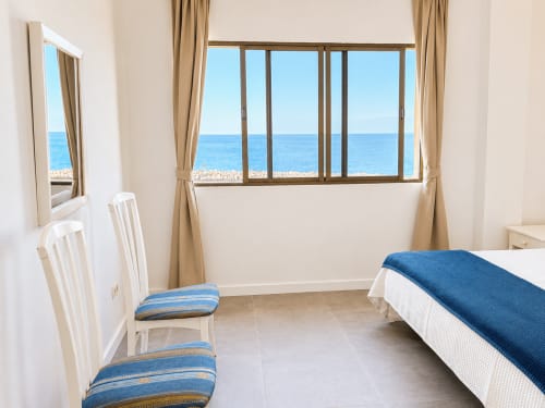 Dormitorio costero luminoso en Tenerife con vistas al mar, sillas blancas, cojines azules y manta azul en la cama