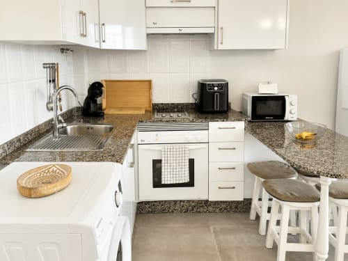 Cocina moderna con armarios blancos, encimeras de granito, electrodomésticos de acero inoxidable y zona de comedor en apartamento de Tenerife