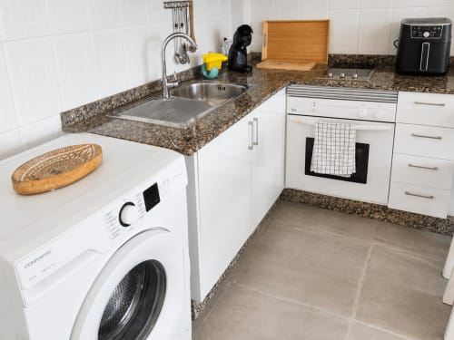 Cocina moderna con fregadero de acero inoxidable, armarios blancos, encimera de granito, lavadora y electrodomésticos en Tenerife