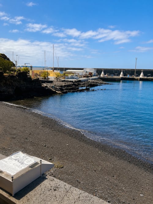 Marina costera en Santa Cruz de Tenerife con veleros, costa rocosa, playa de arena y agua azul en un día soleado
