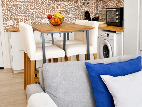 Moderna cocina-comedor con mesa de madera, sillas crema, sofá gris con cojín azul y electrodomésticos en Santa
