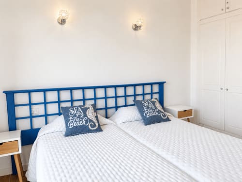 Luminoso dormitorio con cabecero azul de madera, ropa de cama blanca y cojines náuticos en apartamento frente al mar de Tenerife