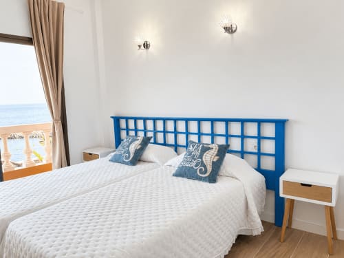 Luminoso dormitorio costero con cabecero azul, ropa de cama blanca, vista al mar y decoración moderna en apartamento de Tenerife