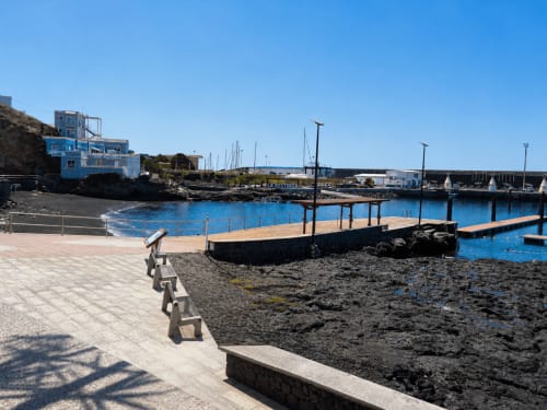 Paseo costero con muelle de madera, aguas azules y edificio de apartamentos azul en Restinga La Playa, Tenerife