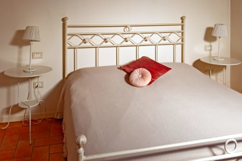 Quarto elegante na Corte delle Giuggiole com cama de latão, roupa de cama creme, almofada vermelha e mesas laterais com luminárias em Cologna