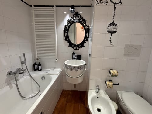 Bagno moderno dell'appartamento Domus Caecilia Roma con specchio nero ornamentale, lavabo tondo, vasca, bidet e pavimenti in legno.