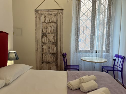 Camera da letto dell'appartamento Domus Caecilia a Roma con letto, asciugamani arrotolati, sedie viola, pannello decorativo in legno e finestra