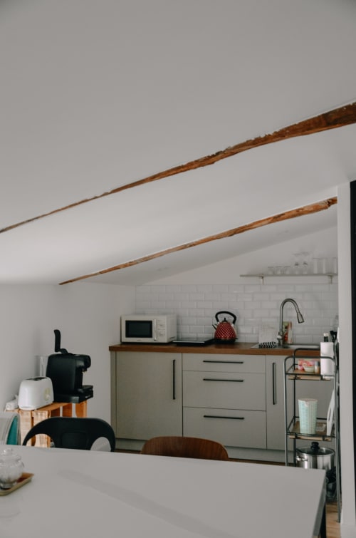 Modern kitchenette in La Maison d'Augustin apartment with white subway tile, wooden beams, and sage cabinetry in