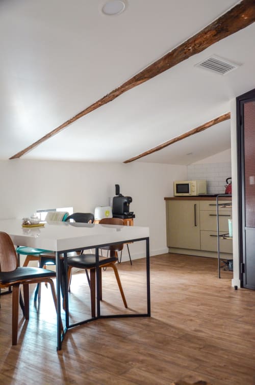 Modern attic apartment with exposed wooden beams, white ceiling, wooden floors, dining table with chairs, kitchenette, and