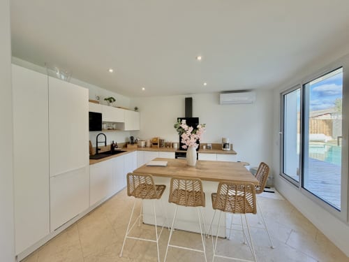 Cuisine moderne avec armoires blanches, table en bois, chaises en rotin, vue sur piscine à la villa LOCATION COTE LANDES