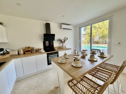 Cuisine moderne de villa avec salle à manger surplombant la piscine, placards blancs, comptoirs en bois et chaises en rotin à Côte Landes.