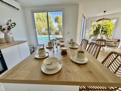 Salle à manger moderne de la villa Pontenx avec vue sur la piscine, table en bois dressée, chaises en rotin et accès à la cuisine.