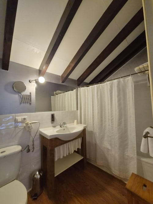 Bathroom with wooden beams ceiling, white sink, toilet, and shower curtain at Posada Torre del Milano, Comillas