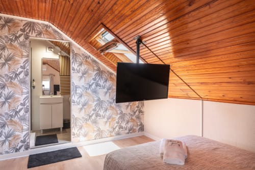 Modern attic bedroom at Charme da Graça, Costa de Caparica, with wooden ceiling, TV, ensuite bathroom, and floral wallpaper