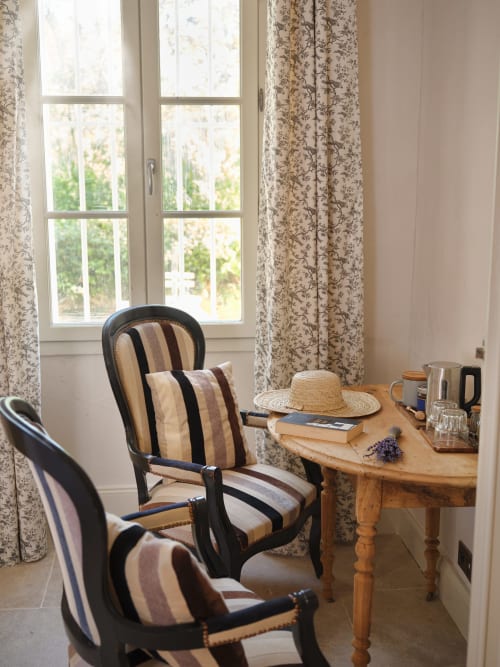 Coin de lecture douillet à la Ferme Saint-Georges avec chaises rayées, bureau en bois, vue sur le jardin et rideaux à fleurs