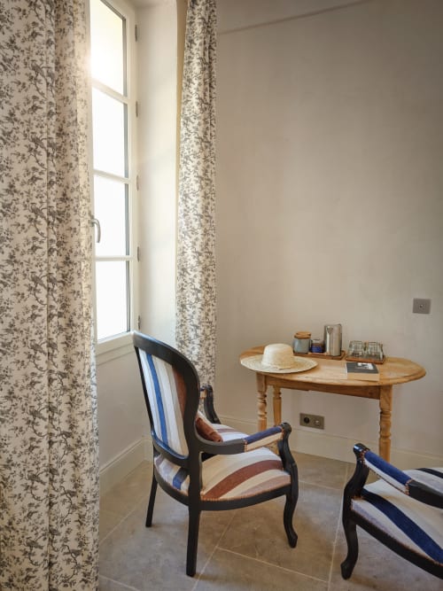 Coin cosy à la Ferme Saint-Georges avec rideaux fleuris, bureau en bois, chaise rayée et lumière naturelle