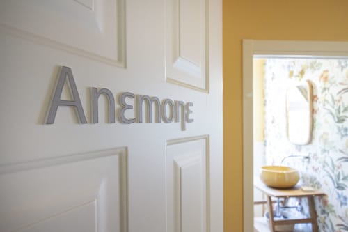 Ingresso della camera Anemone presso l'Epimoni B&B di Catania, con scritte grigie su parete bianca e scorcio dell'interno dai toni giallastri