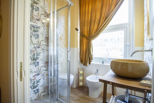 Lussuoso bagno nella suite Anemone di Epimoni B&B, Catania. Lavabo in ceramica dorata, doccia piastrellata, finestra con vista città e tende.