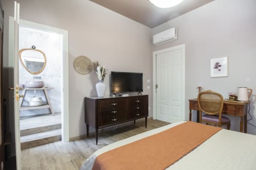 Camera da letto Epimoni B&B a Catania con letto, cassettone in legno scuro, TV, scrivania e bagno en-suite visibile dalla porta aperta