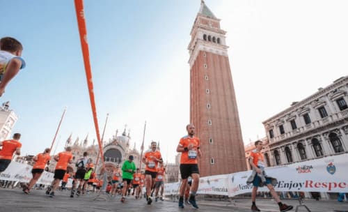 Maratoneti in magliette arancioni corrono attraverso la Piazza San Marco di Venezia con l'iconico campanile sullo sfondo