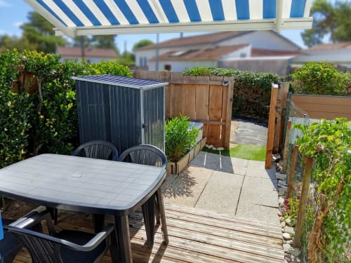 Terrasse ensoleillée aux Relais-Orbigny avec table de salle à manger, auvent rayé bleu, abri de jardin et haies luxuriantes