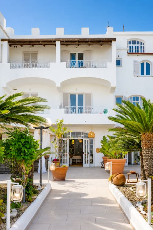 Cortile del Villa Bina Sea Hotel con architettura mediterranea bianca, palme e vasi in terracotta a Serrara Fontana