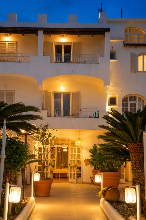 Ingresso del cortile illuminato del Villa Bina Sea Hotel al tramonto con architettura bianca e palme, Serrara Fontana