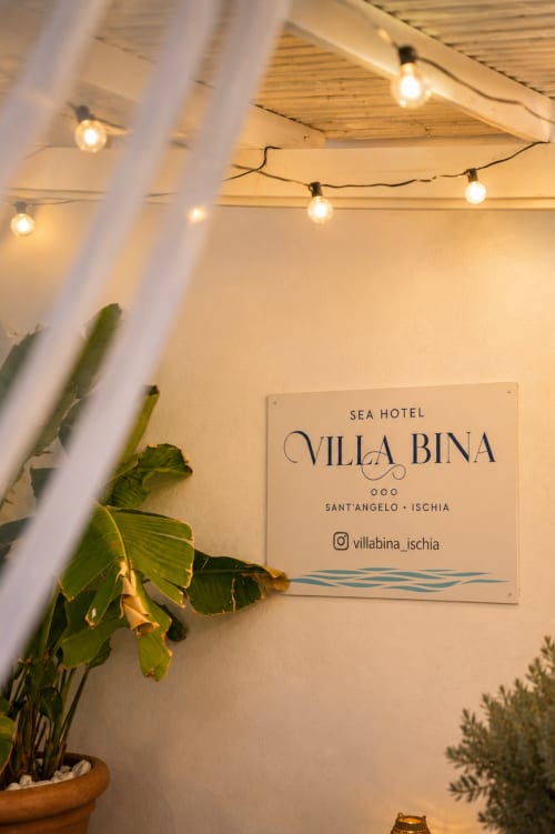 Insegna del Villa Bina Sea Hotel illuminata da calde luci decorative e circondata da piante tropicali a Sant'Angelo, Ischia