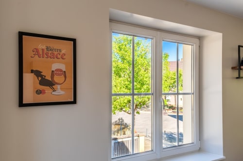 Affiche de bière d'Alsace encadrée sur mur beige à côté de grandes fenêtres donnant sur les arbres verts et la cour de l'hôtel Les Ambrades