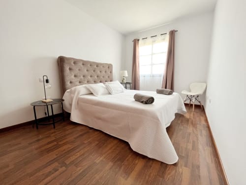 Dormitorio luminoso en villa Casa Laferre, La Oliva, con cabecero capitoné, ropa de cama blanca, suelos de madera y grandes ventanales