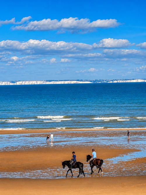 Twee ruiters te paard die het zandstrand van Ô de Mer, Sangatte oversteken, met witte kliffen en blauwe zee op de achtergrond