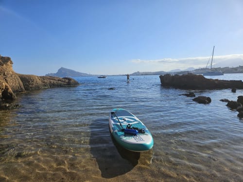 Planche de paddle turquoise dans les eaux peu profondes de la Méditerranée près d'Altea, Alicante, avec une côte rocheuse et des voiliers.