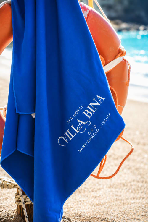 Asciugamano blu con il marchio di Villa Bina Sea Hotel sulla spiaggia sabbiosa di Ischia, Italia