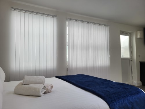 Bright studio bedroom at Cosy in Cork with white vertical blinds, white bedding, rolled towels, and dark blue throw blanket