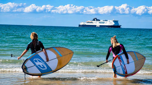 Two windsurfers in wetsuits holding boards at Ô de Mer beach in Sangatte with ferry ship offshore.