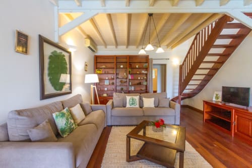 Grand salon/TV avec canapés. Architecture  créole avec poutres en bois, meubles et parquet en teck. Escalier permettant d'accéder à la chambre quadruple à l'étage.