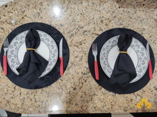 Dois aparelhos de mesa com guardanapos pretos, pratos estampados e talheres com cabo vermelho sobre bancada de granito no Wellington Rodovalho
