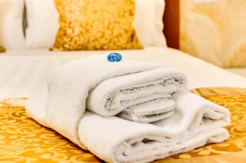 Asciugamani bianchi arrotolati con sigillo blu dell'hotel su un letto con motivi dorati nella Deluxe Queen Room Domus Praeclara Merulana